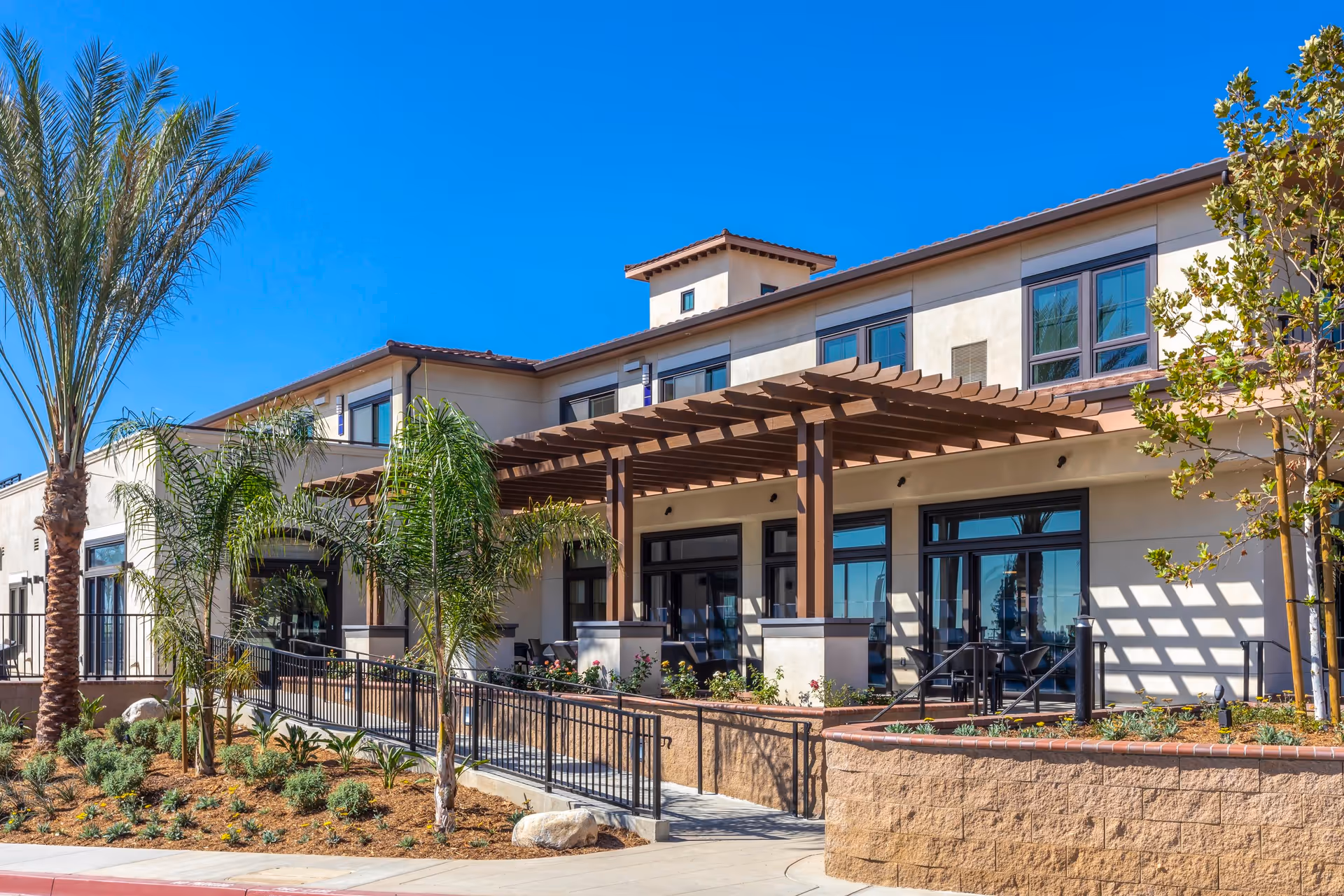 Exterior view of a modern two-story building with large windows and a wooden pergola over a patio area. The building is surrounded by landscaped plants, palm trees, and a ramp with a black metal railing. The sky is clear and blue.