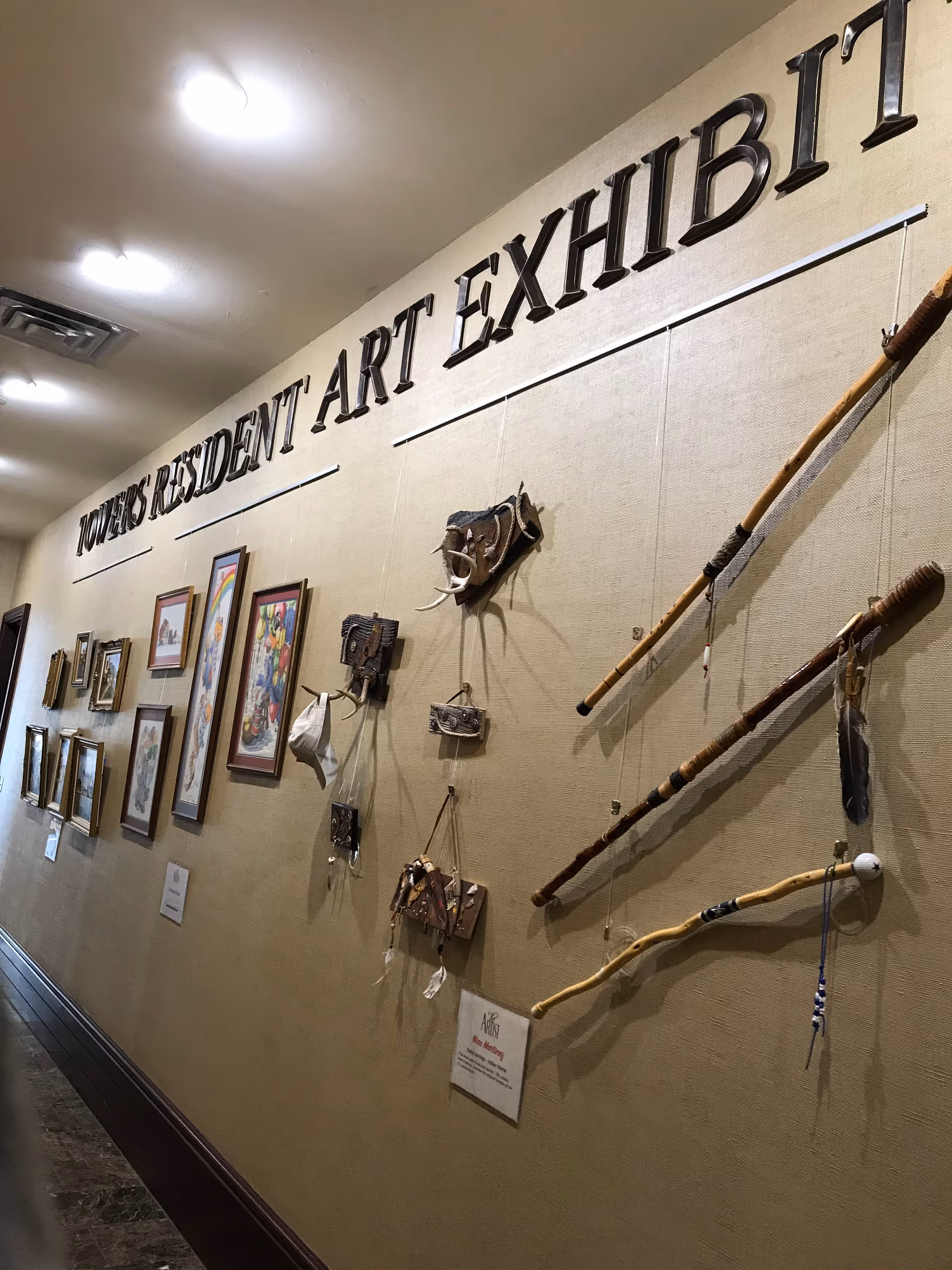 A hallway wall displaying various framed artworks and handcrafted items as part of the Towers Resident Art Exhibit. The wall has a beige fabric covering and the exhibit title is mounted in large black letters above the artwork.