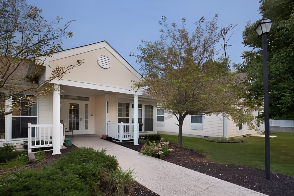 Exterior view of a single-story building with a covered entrance supported by white columns, a paved walkway leading to the entrance, surrounded by green shrubs, trees, and a lawn under a clear blue sky.