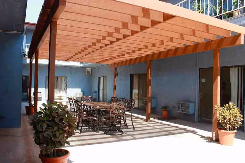 Shaded outdoor patio with a wooden pergola, metal dining table and chairs, potted plants, and blue building walls.