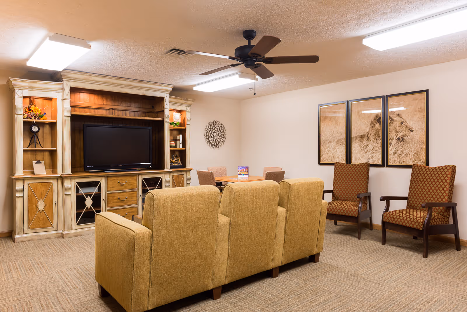 A cozy living room area in an assisted living center featuring a beige couch facing a wooden entertainment center with a flat-screen TV. The room also has two patterned armchairs, a ceiling fan, and framed artwork of a lion on the wall.