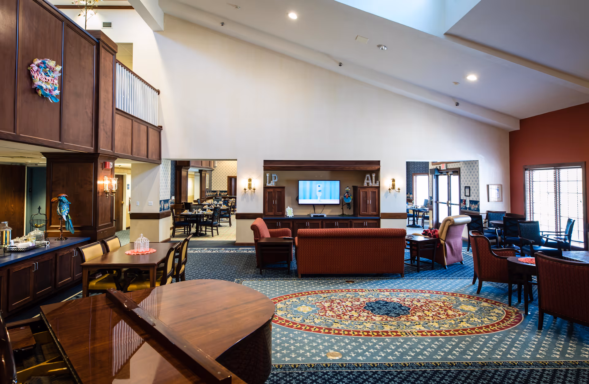 Spacious assisted living facility common area with high ceilings, featuring a grand piano in the foreground, multiple seating arrangements including sofas and armchairs, a wall-mounted TV, and a dining area visible through an open doorway. The room has patterned carpeting, wood paneling, and large windows allowing natural light.