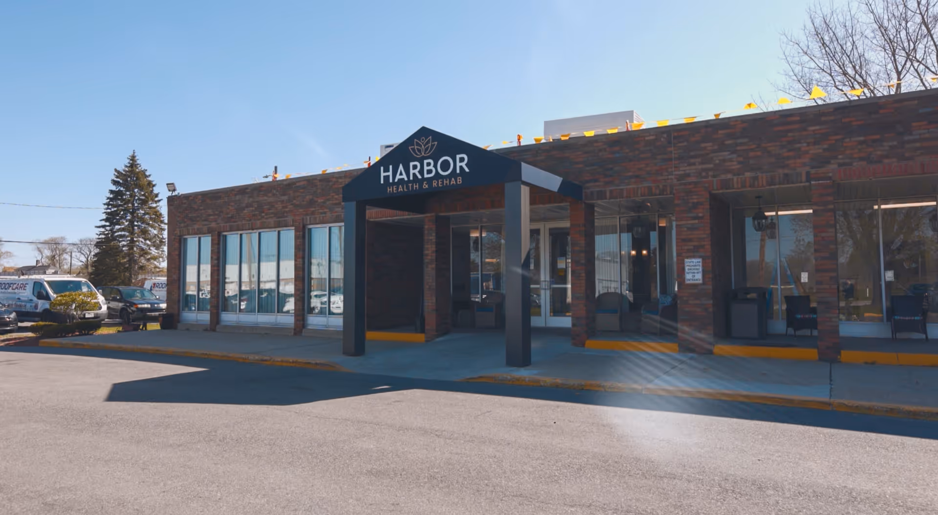 Exterior view of Harbor Health & Rehab building with a brick facade, large windows, and a covered entrance. Several parked vehicles are visible to the left, and a clear blue sky is overhead.
