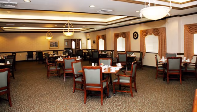 A spacious dining room with multiple wooden tables and chairs arranged neatly. Each table is set with white cups, plates, and condiments. The room has beige walls with decorative curtains on the windows and ceiling lights providing warm illumination.