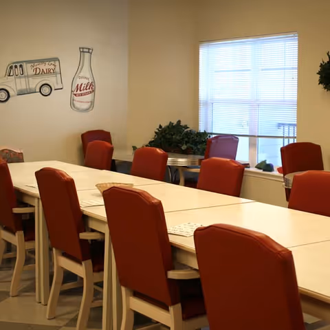 A dining room with a long white table surrounded by red cushioned chairs. There is a window with blinds letting in natural light, a small round table with plants near the window, and wall decorations including a vintage-style milk bottle and delivery truck.