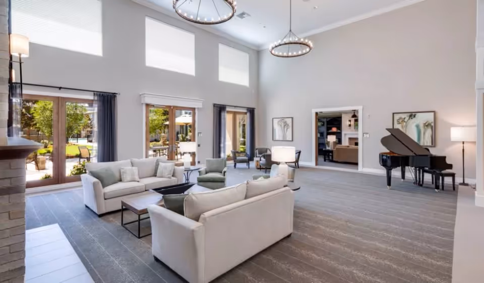A spacious and well-lit living room area with two beige sofas, a green armchair, and a coffee table in the center. Large windows and glass doors allow natural light to fill the room, showing an outdoor patio with seating. The room features high ceilings with two circular chandeliers, a grand piano on the right side, and framed artwork on the walls. The floor is carpeted in a neutral tone, and there are additional chairs near the windows.