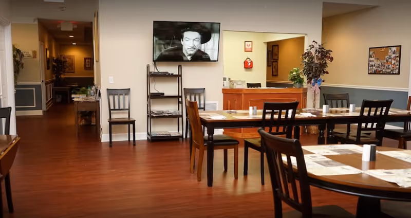 Interior view of a dining area in a senior living facility with wooden tables and chairs arranged neatly. A television mounted on the wall displays a black and white movie. There is a small reception or service counter in the background with plants and decorations, and a hallway leading to other rooms is visible on the left side.