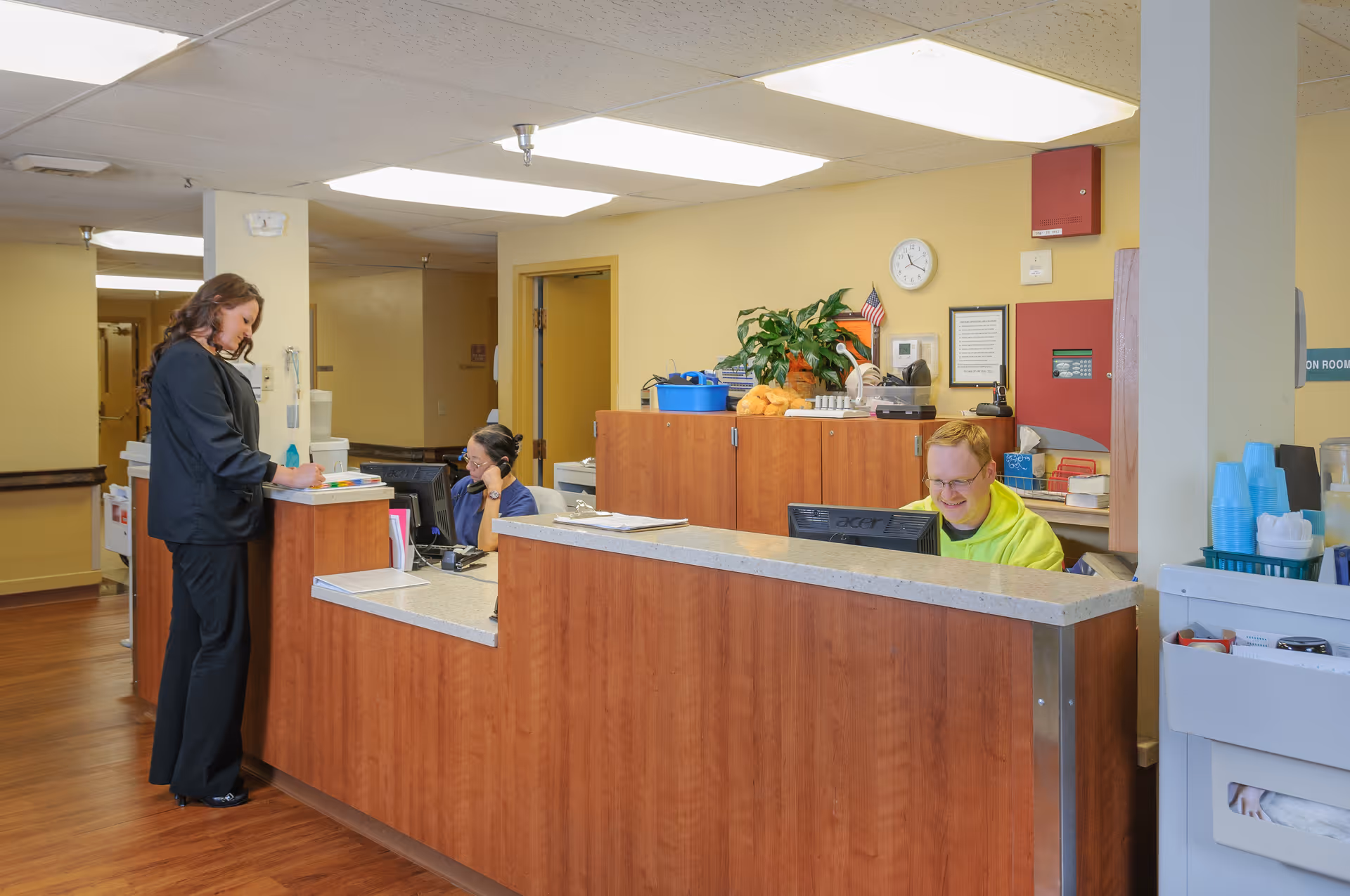 Reception area in a senior living facility with two staff members behind a wooden desk. One staff member is seated and talking on the phone, while the other is working on a computer. A woman is standing at the desk, writing on a clipboard. The area has yellow walls, overhead fluorescent lighting, and wooden flooring. There are plants, office supplies, and a clock on the wall behind the desk.