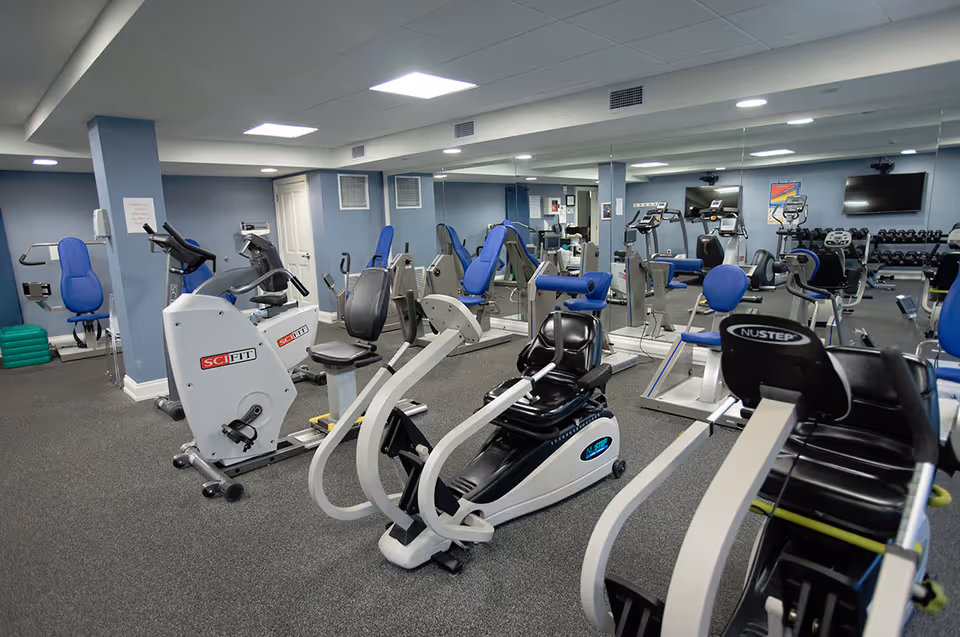 Interior view of a fitness room with various exercise equipment including stationary bikes, weight machines, and a rack of dumbbells. The room has blue walls, a gray carpeted floor, and a large mirror covering one wall.