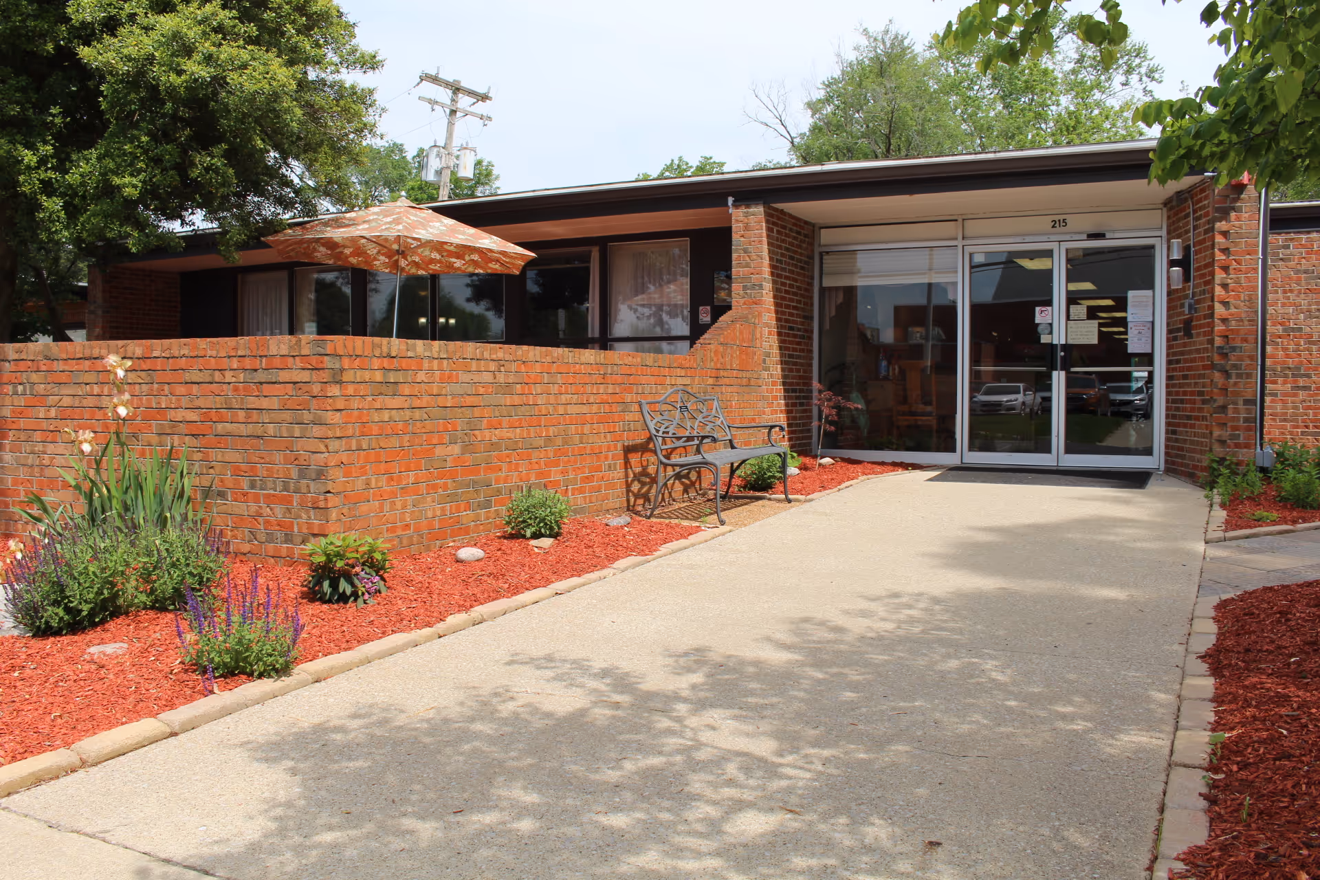 Entrance to a brick building with a glass door marked 215, a concrete pathway leading to the door, a metal bench on the right side, and landscaped flower beds with red mulch and various plants on both sides. There is also a patio area with an orange umbrella visible behind a brick wall.