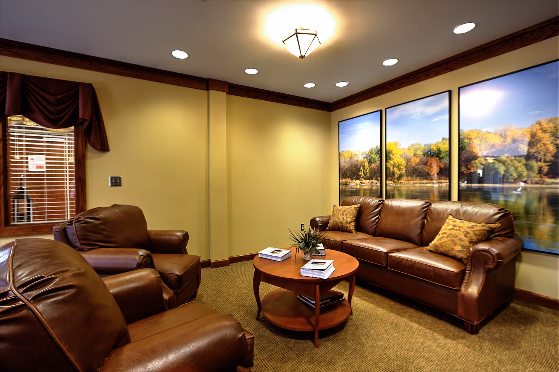 A cozy living room with brown leather sofa and two matching armchairs arranged around a wooden coffee table with books and a small plant. The walls are painted yellow with dark wood trim, and three large framed pictures of a lake and autumn trees hang on one wall. A window with wooden blinds and a dark red valance is visible on the left side.