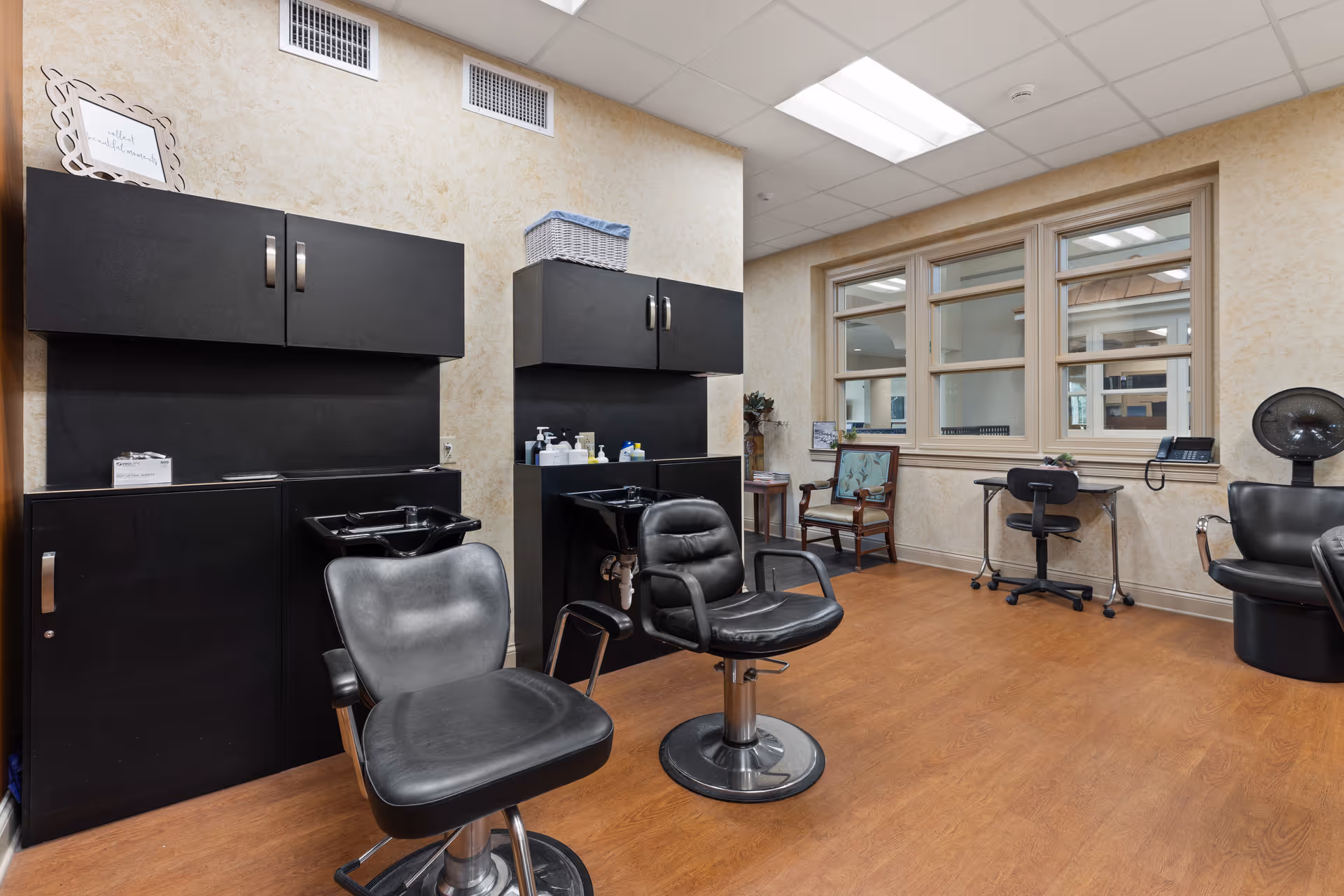 Interior salon room with black styling chairs and wash basins, cabinets, and seating by a window.