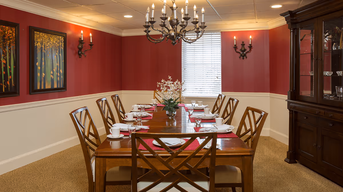 A formal dining room with a long wooden table set for eight people. The table has white plates, cups, glasses, and red placemats. There is a floral centerpiece in the middle of the table. The walls are painted red with white wainscoting, decorated with two framed paintings and wall sconces with candles. A chandelier hangs above the table, and a large wooden china cabinet is on the right side of the room.