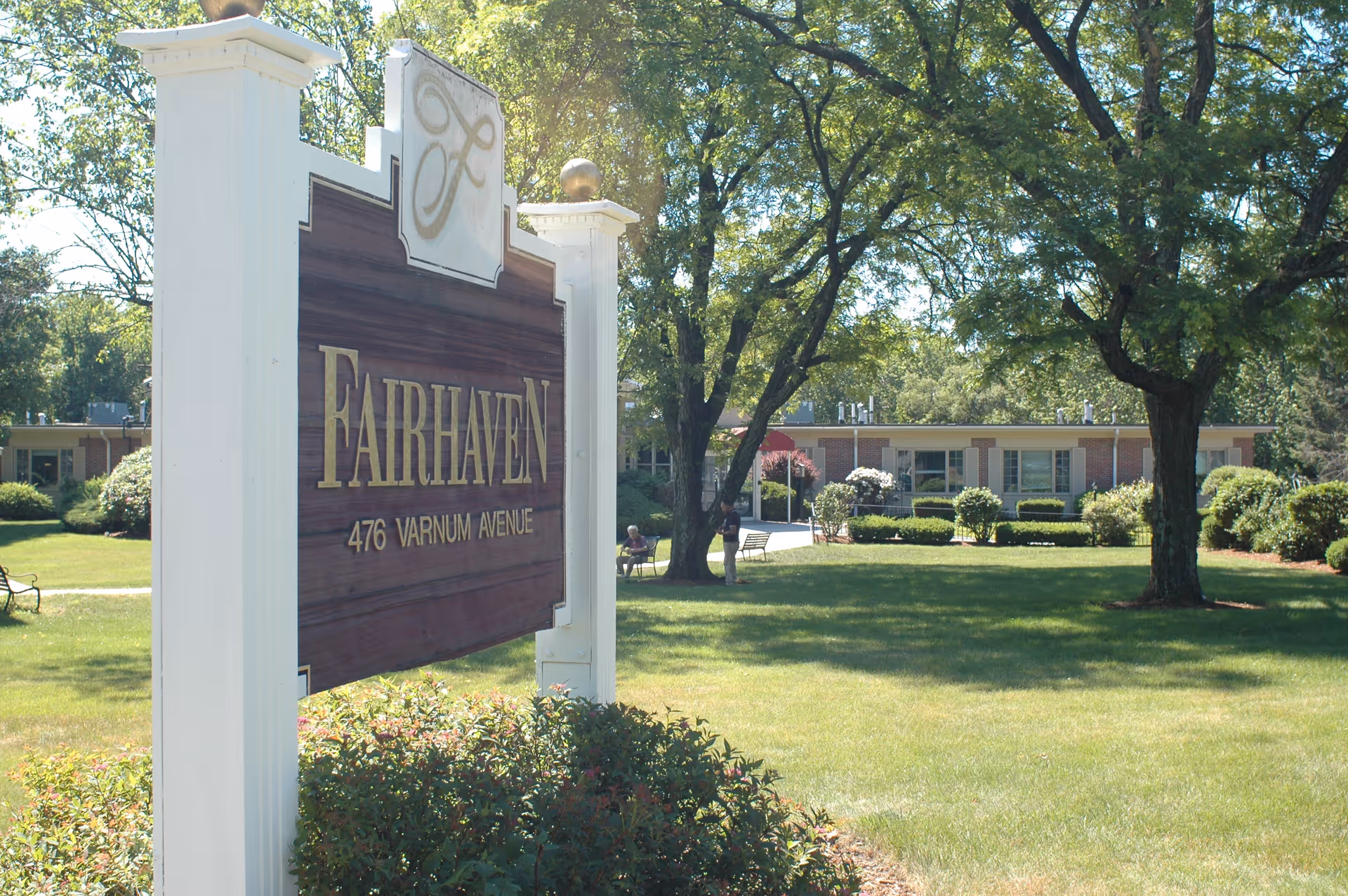 Outdoor view of Fairhaven Healthcare Center for Rehabilitation and Nursing sign with the address 476 Varnum Avenue. The sign is white with gold lettering and stands in front of a green lawn with trees and a building in the background.
