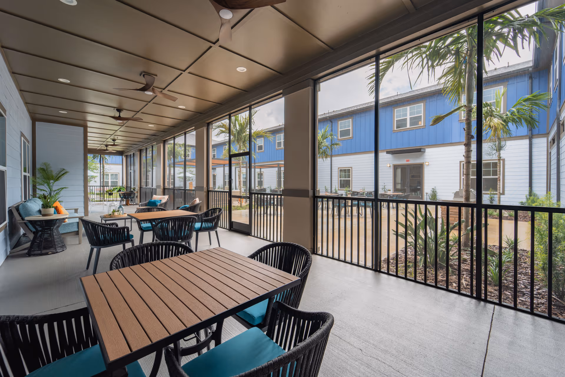 A covered outdoor patio area with multiple tables and chairs, ceiling fans, and potted plants. The patio is enclosed with large screened windows, overlooking a courtyard with palm trees and a two-story building painted blue and white in the background.