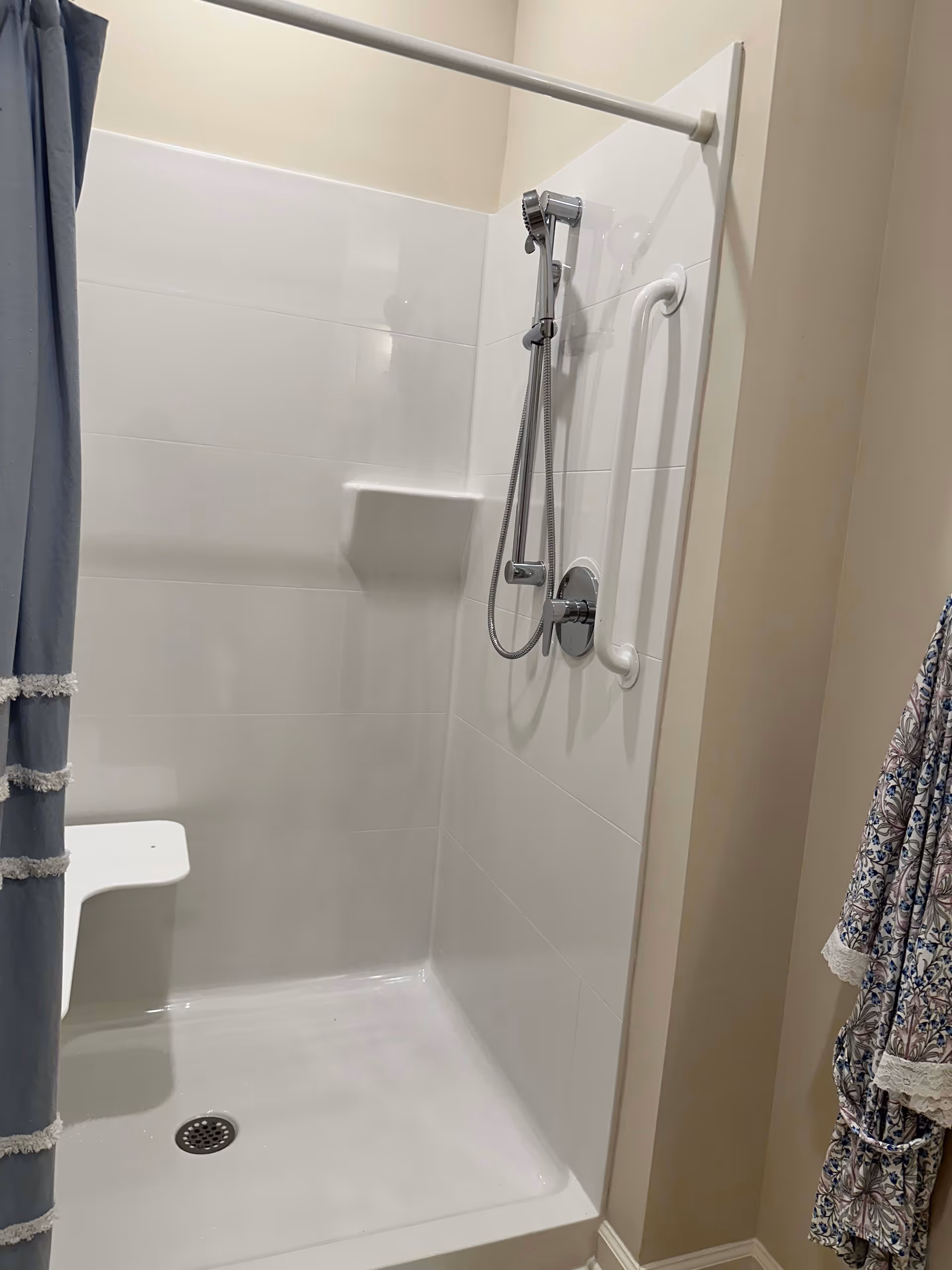 A clean, white tiled shower stall with a handheld showerhead mounted on the wall, a built-in corner shelf, a white grab bar, and a small white fold-down shower seat. A blue shower curtain with white tassels is partially visible on the left, and a patterned bathrobe hangs on the right wall outside the shower.
