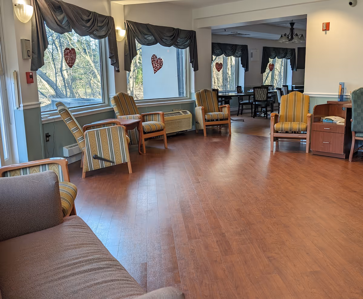 A spacious senior living facility common area with wooden flooring, several striped armchairs arranged along the walls, large windows with dark valances, and heart-shaped decorations hanging on the windows. In the background, there is a dining area with tables and chairs.