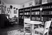Small community interior with bookshelves along the wall, a round table and chairs, and a piano.