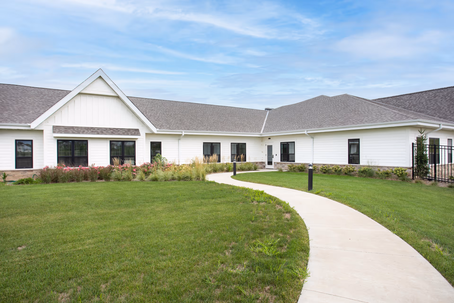 Exterior view of a single-story white senior living facility building with a gray roof, multiple windows, a curved concrete walkway leading to the entrance, green lawn, and landscaping with shrubs and flowers under a partly cloudy sky.