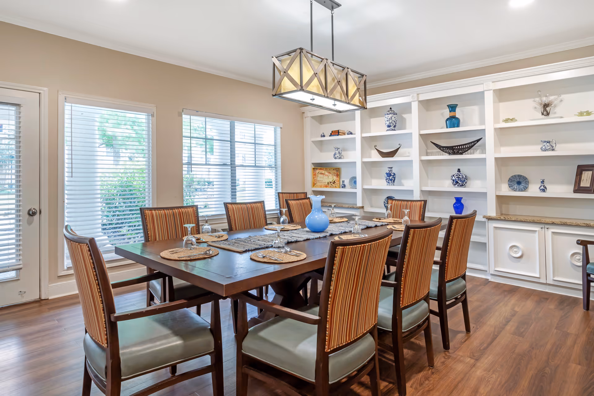 A dining room with a large wooden table set for eight people with placemats, upside-down wine glasses, and a blue vase centerpiece. The room has wooden flooring, large windows with blinds, a door to the left, and white built-in shelves decorated with various blue and white vases and decorative items. A modern rectangular chandelier hangs above the table.