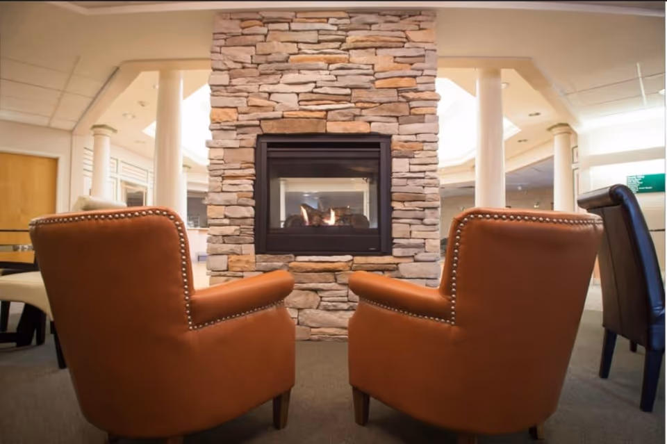Two brown leather armchairs facing a stone fireplace with a fire burning inside, situated in a well-lit room with white columns and additional seating visible in the background.