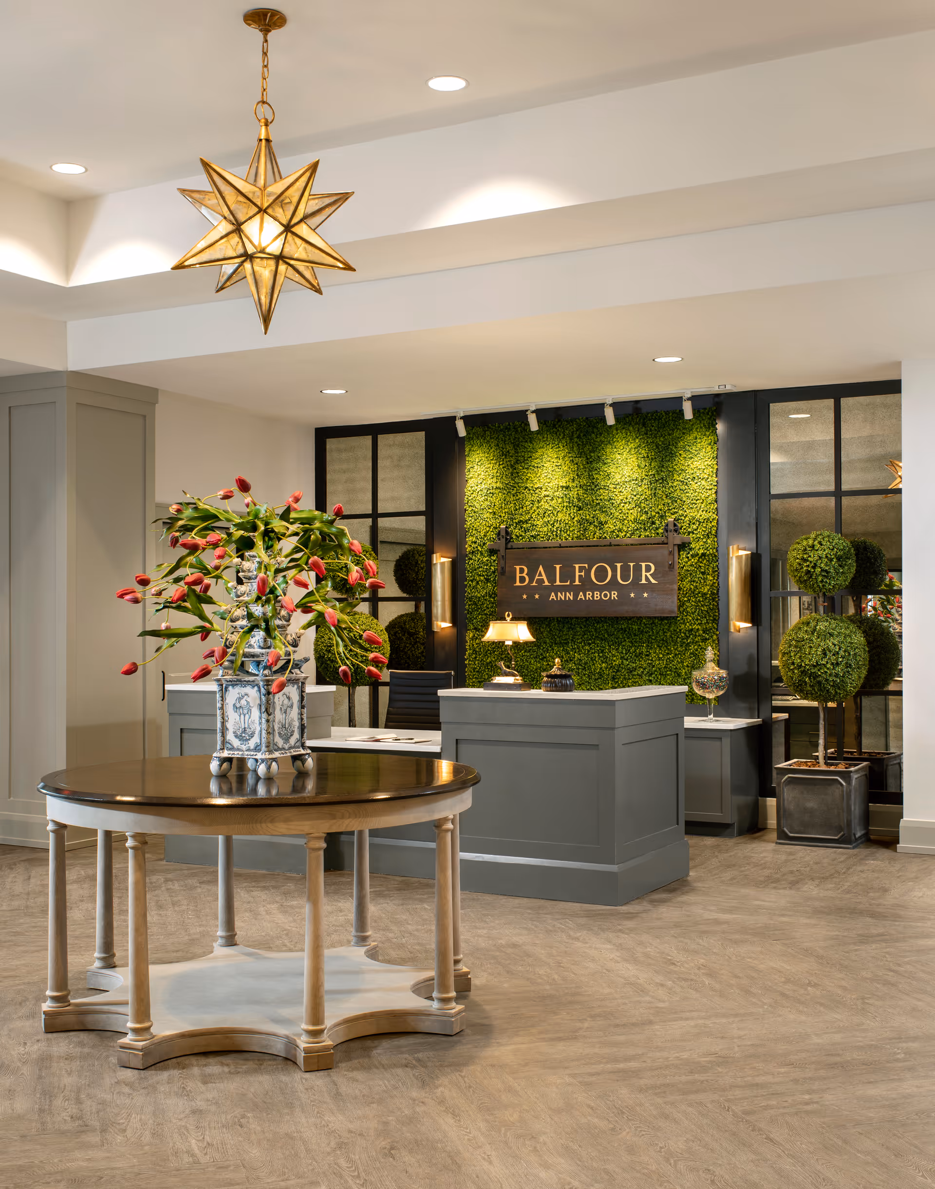 Reception area with a round wooden table holding a vase of red tulips in the foreground. Behind the table is a gray reception desk with a lamp and decorative items. The back wall features a green plant wall with a wooden sign that reads 'BALFOUR ANN ARBOR'. Two topiary plants are placed on the right side of the desk. The ceiling has recessed lighting and a decorative star-shaped pendant light.