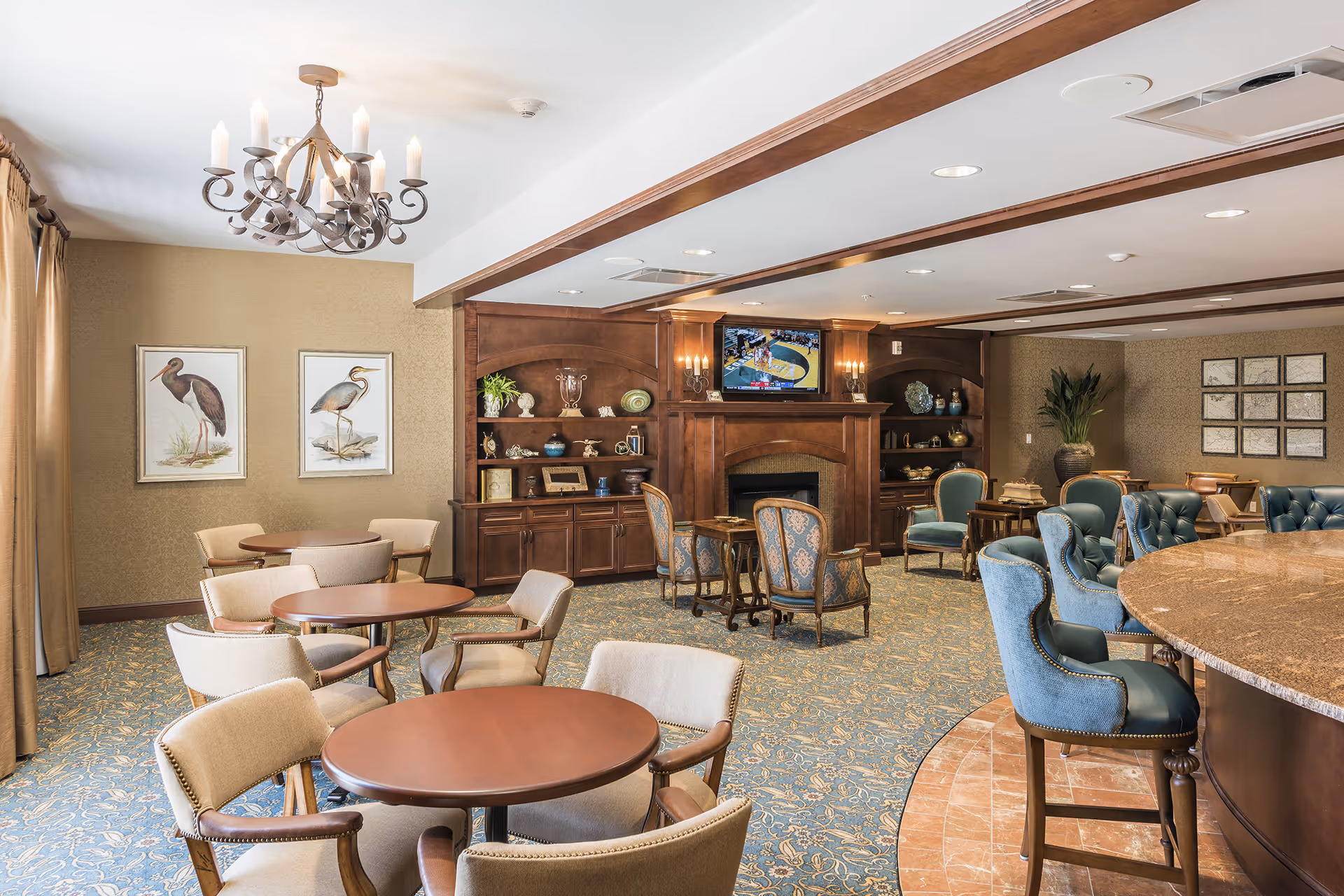 A spacious and elegantly furnished common area with round wooden tables and cushioned chairs, a large wooden fireplace with built-in shelves displaying decorative items, a mounted TV above the fireplace, and a chandelier hanging from the ceiling. The room has patterned carpet flooring and beige walls adorned with framed bird artwork and plants.