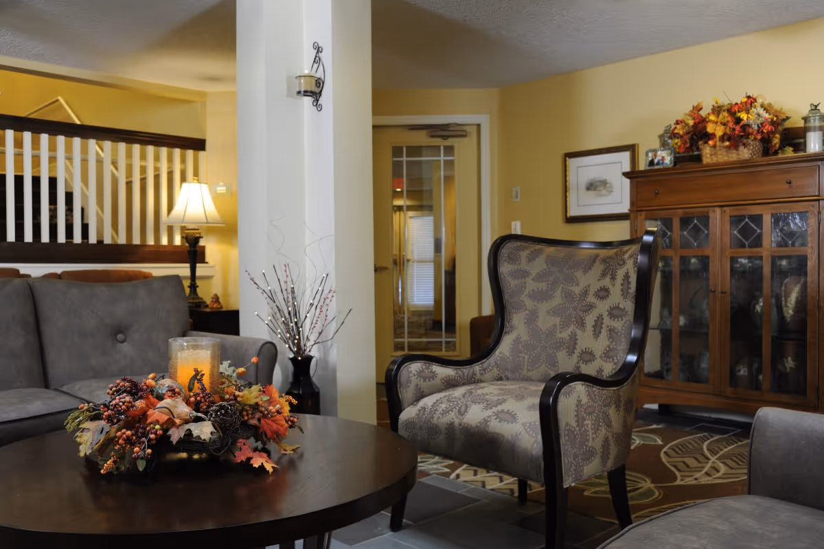 Seating area with a patterned armchair and gray sofa around a round coffee table topped with a fall floral centerpiece, beside a wooden display cabinet.