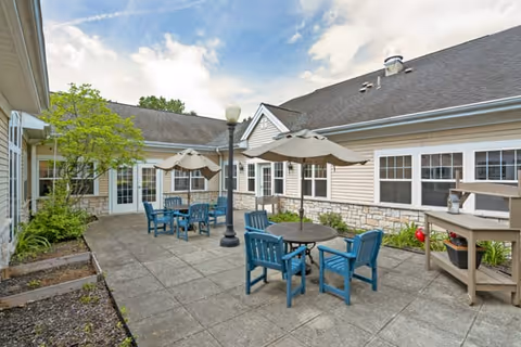 Outdoor patio area at Brookdale East Niskayuna with several blue chairs and round tables shaded by beige umbrellas. The patio is paved with concrete tiles and surrounded by beige siding buildings with white-trimmed windows. There is a lamppost in the center and some greenery along the edges.