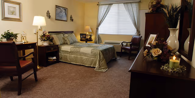 A cozy bedroom with a neatly made bed covered in a green quilt. The room features a wooden nightstand with a lamp and decorative items, a wooden dresser with flowers and a lit candle, two chairs, a window with blinds and curtains, and soft carpeting.