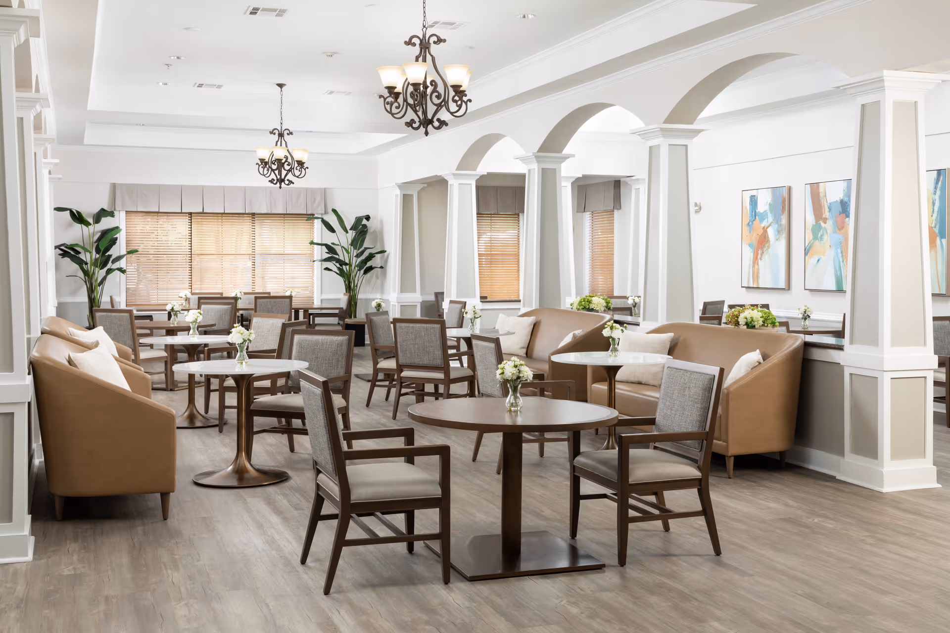 Bright and spacious dining room with multiple round tables and cushioned chairs arranged neatly. The room features beige leather sofas along the walls, decorative plants, large windows with wooden blinds, white walls with arches and columns, and modern chandeliers hanging from the ceiling. Abstract paintings adorn the walls, and small flower vases are placed on each table.