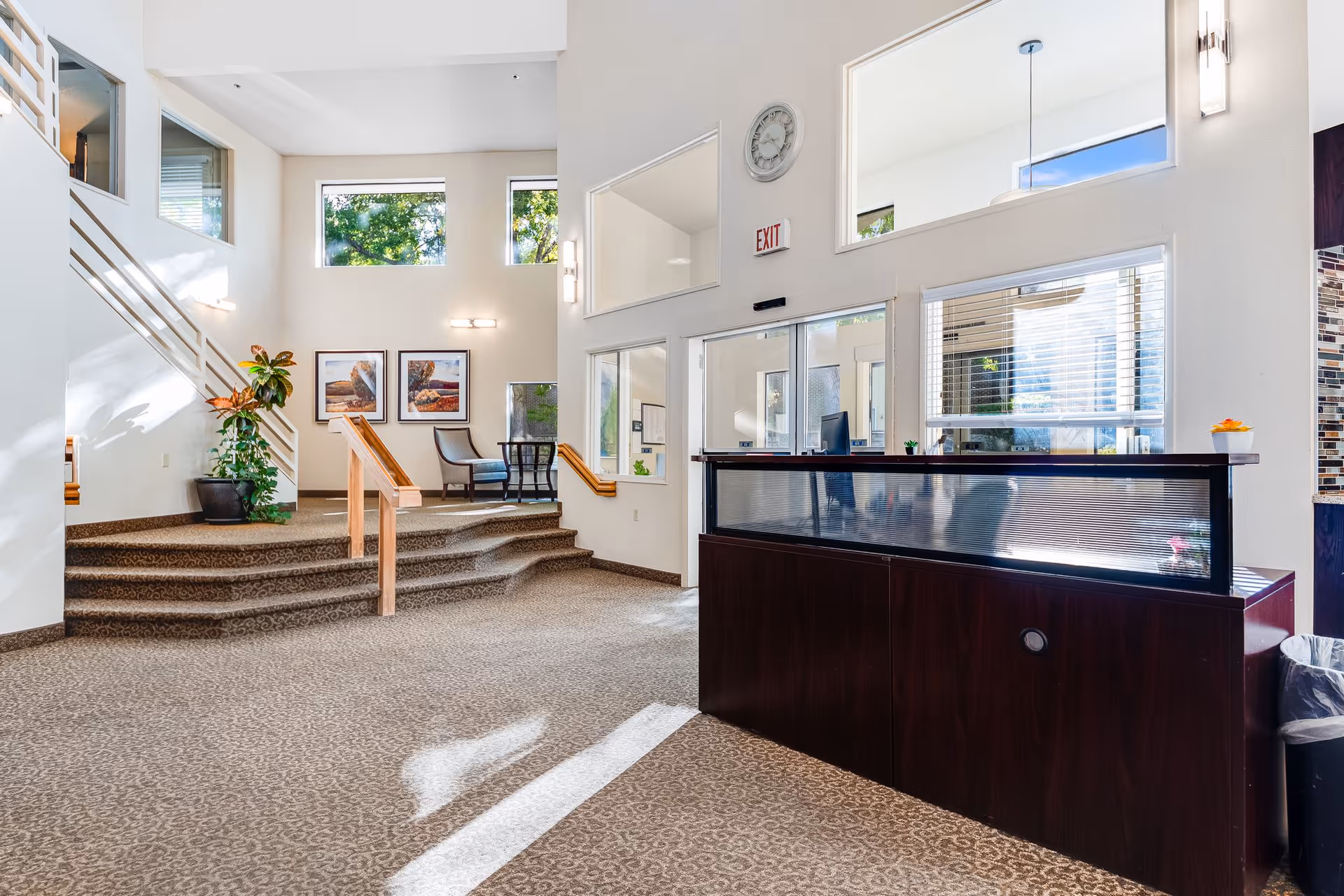 Bright and spacious reception area with a dark wood front desk on the right, carpeted floor, stairs with wooden handrails leading to a seating area with two chairs and a small table, large windows letting in natural light, and framed artwork on the wall.