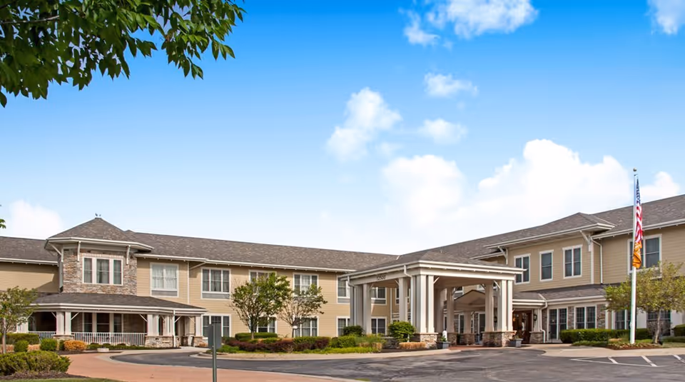 Exterior view of Sunrise of Overland Park senior living facility showing a two-story building with beige siding, stone accents, a covered entrance, landscaped greenery, and an American flag on a flagpole under a blue sky with scattered clouds.