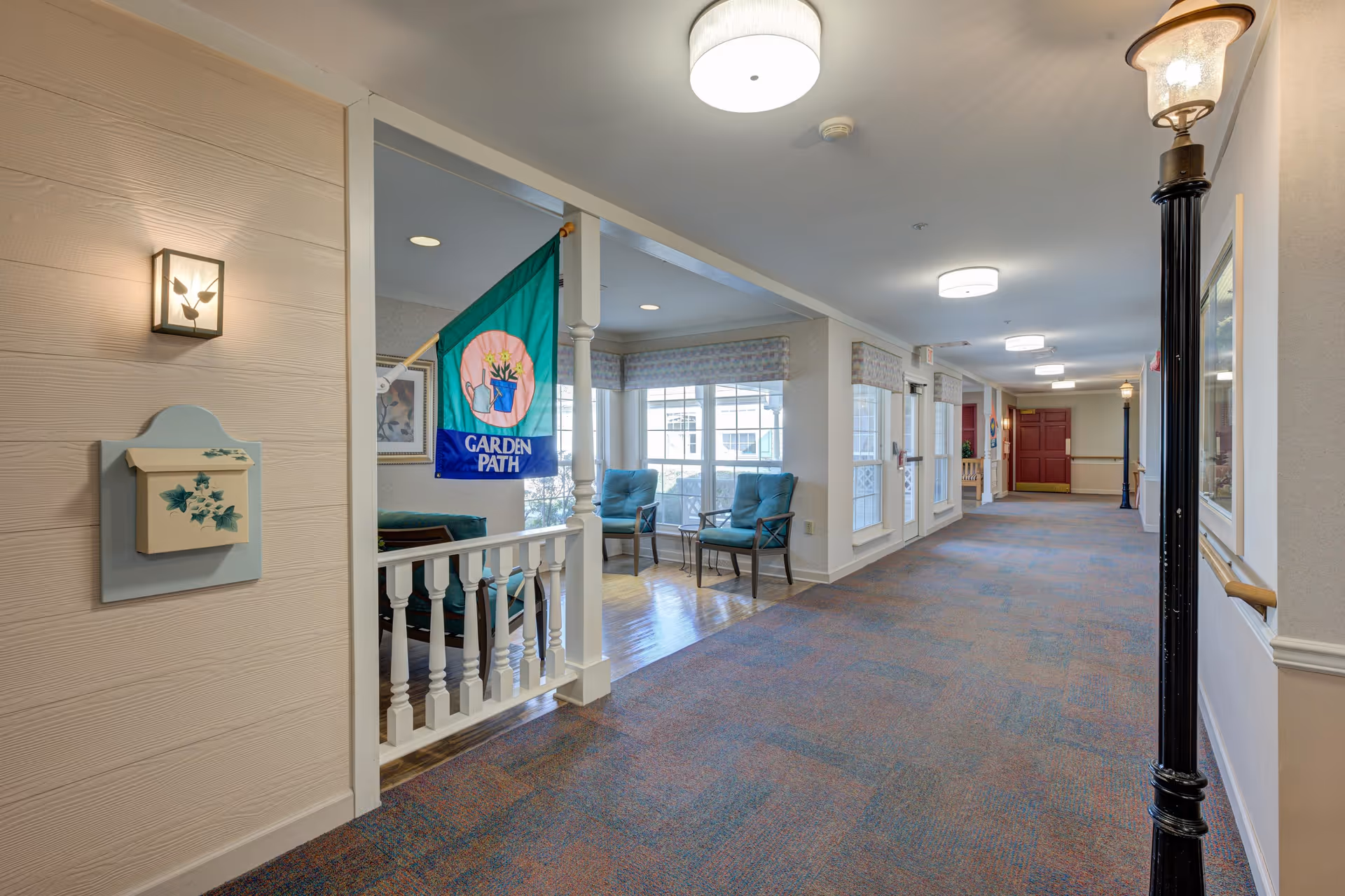 A well-lit hallway in a senior living facility with a carpeted floor and white walls. On the left side, there is a small sitting area with two teal chairs and a small table near large windows. A colorful flag with the words 'Garden Path' hangs near the sitting area. The hallway features decorative streetlamp-style lights and framed artwork on the walls.