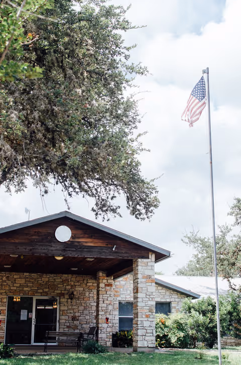 Exterior view of a stone building with a covered entrance and a bench outside. There is a tall flagpole with an American flag flying in front of the building, and large trees surround the area under a partly cloudy sky.