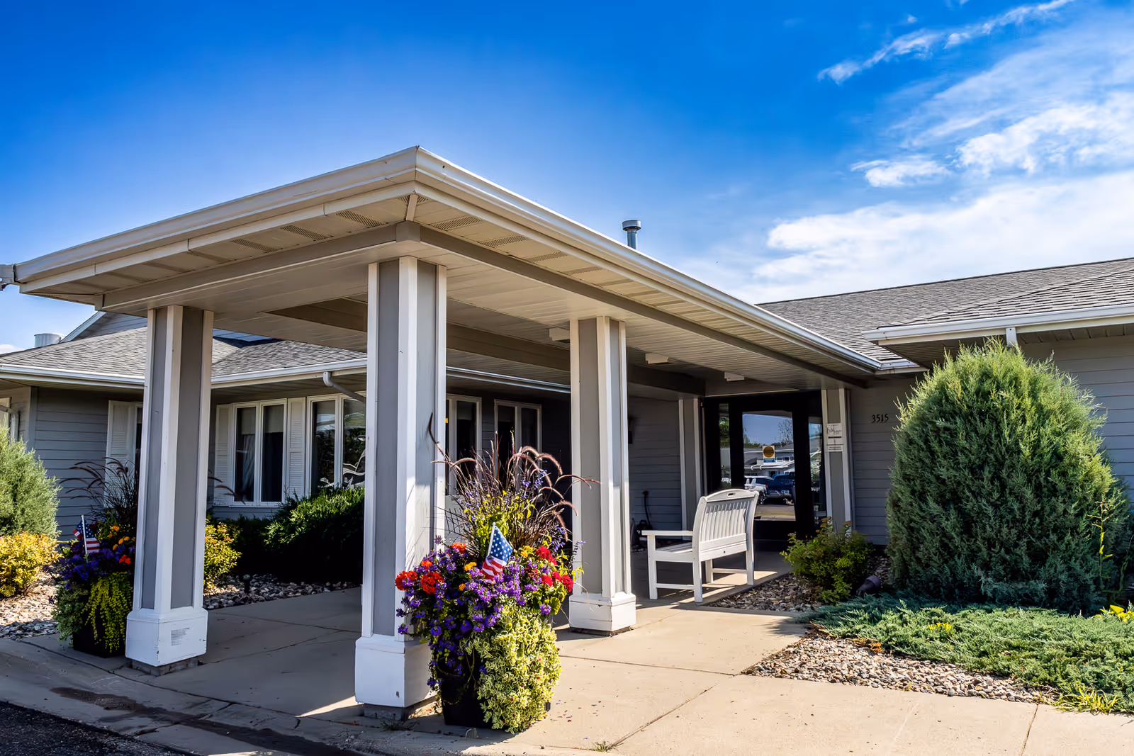 Exterior view of Elison Assisted Living of Minot showing the entrance with a covered porch supported by white columns, a white bench, and large flower pots with colorful flowers and small American flags. The building is light gray with white trim, surrounded by green shrubs and landscaping under a blue sky with some clouds.