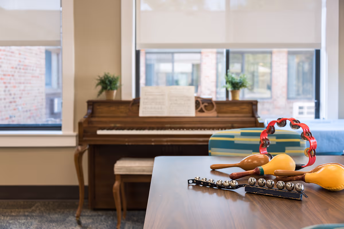 A cozy room with a wooden piano against the wall, two potted plants on top of the piano, and sheet music placed on the piano stand. In the foreground, a table holds various musical instruments including a red tambourine, maracas, and jingle bells. Large windows with beige roller shades allow natural light into the room.