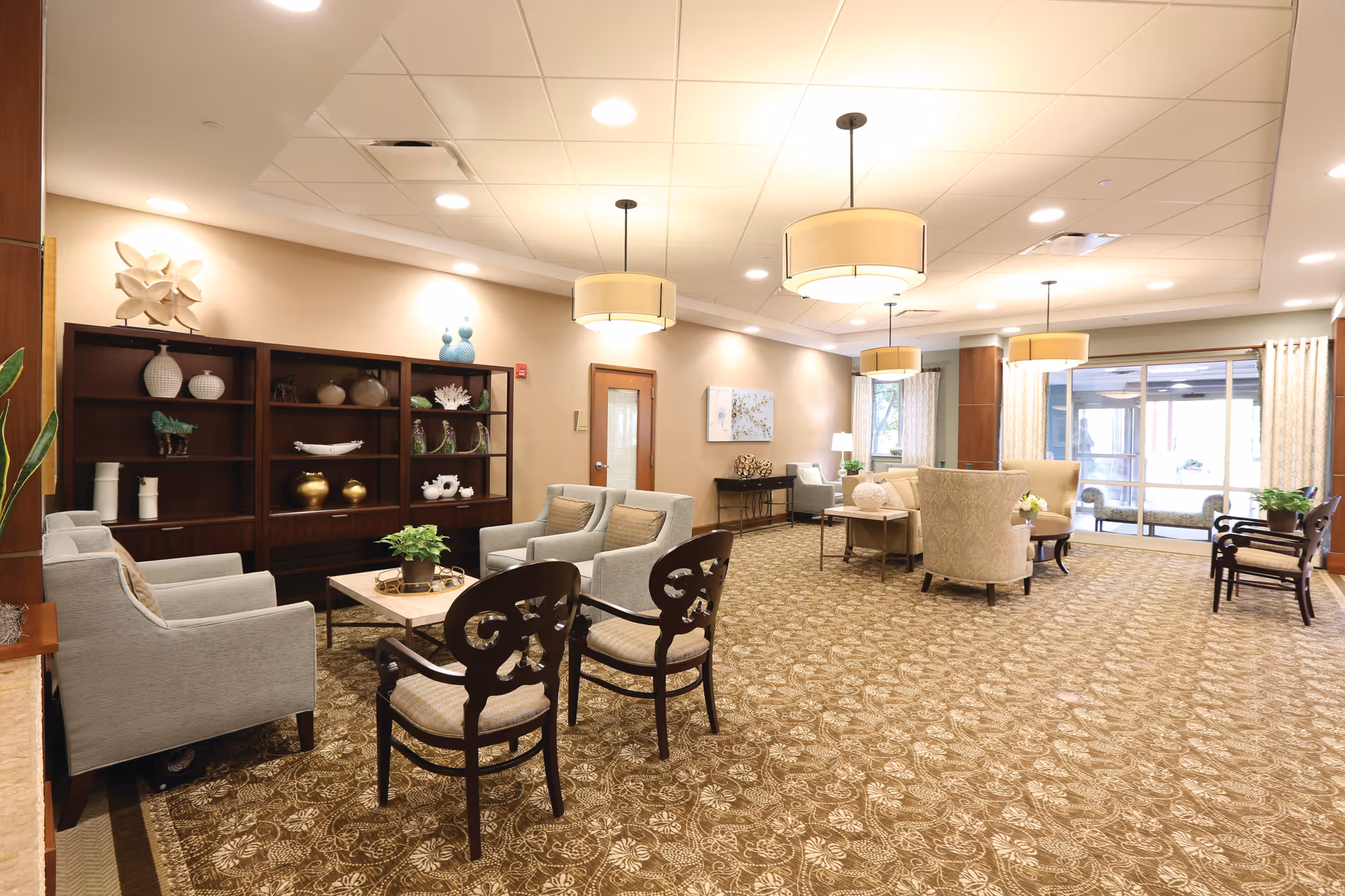 A spacious and well-lit living room area in a senior living facility named Sedgebrook, featuring multiple seating arrangements with armchairs and wooden chairs around coffee tables. The room has a patterned carpet, decorative shelves with vases and ornaments, large windows with curtains, and modern ceiling lights.