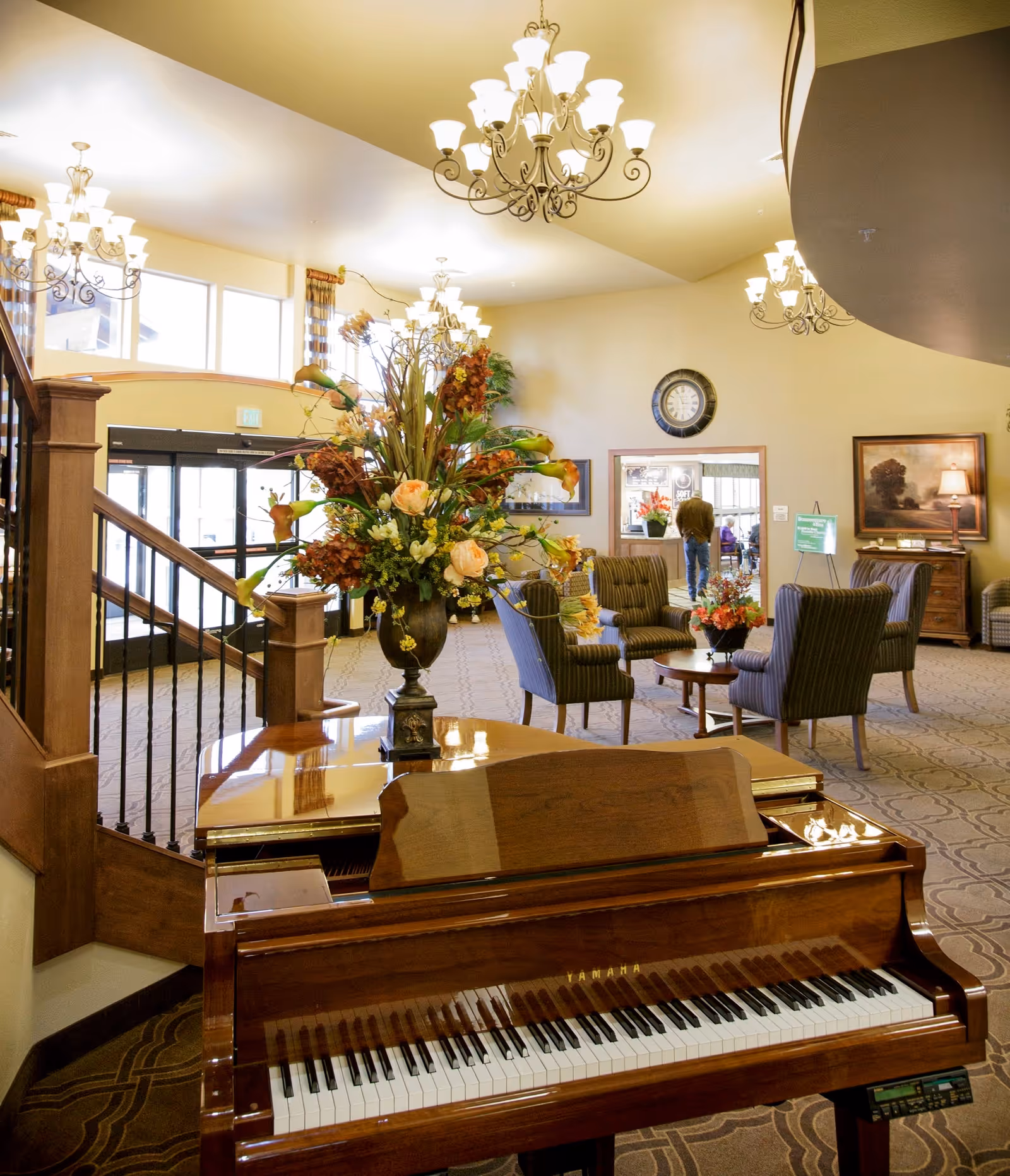 A spacious and well-lit living room area in a senior living facility featuring a polished Yamaha grand piano in the foreground with a large floral arrangement on top. The room has several chandeliers hanging from the ceiling, comfortable armchairs arranged around a small table with flowers, a staircase with wooden railing, and a clock on the wall above a doorway. A person is visible walking in the background near the entrance.