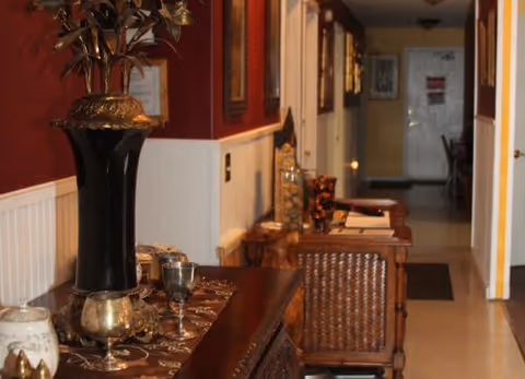 A hallway in a senior living facility with a dark wooden table on the left side adorned with decorative items including a large black vase with a plant, silver goblets, and other small ornaments. The walls are painted red on the upper half and white on the lower half with framed pictures hanging. The hallway extends into the distance with doors and furniture visible along the right side.