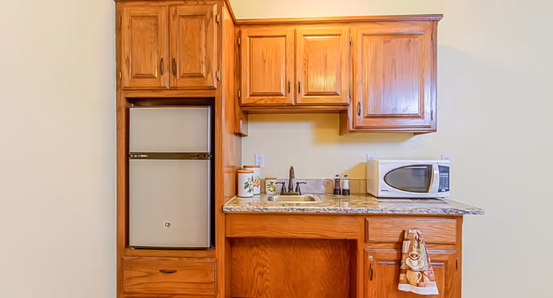 Small kitchenette area with wooden cabinets, a mini refrigerator, a sink with a faucet, a microwave on the countertop, and a decorative towel hanging from a cabinet handle.