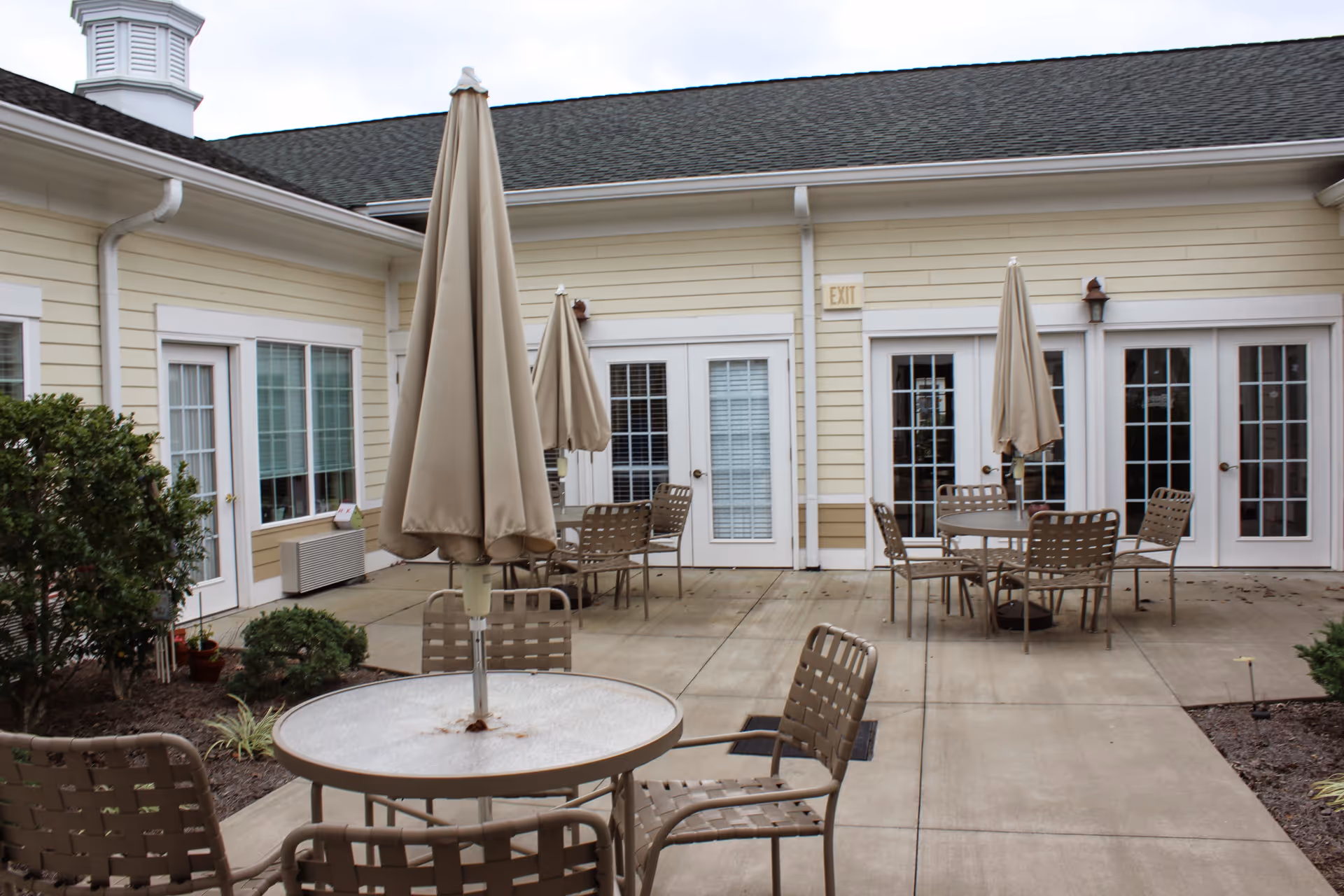 Courtyard patio with round tables, metal chairs, and closed umbrellas outside a pale yellow senior living building.
