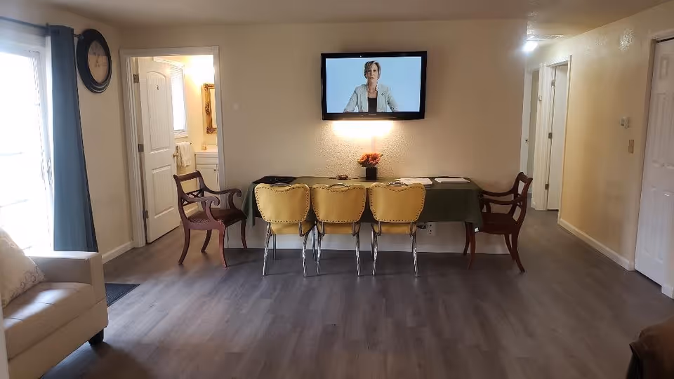 Interior view of a room with a table covered by a green tablecloth, three yellow chairs and two wooden armchairs around it. A flat-screen TV is mounted on the wall above the table displaying a woman. To the left, there is a beige sofa and a large clock on the wall near a window with curtains. Doors and hallway are visible in the background.