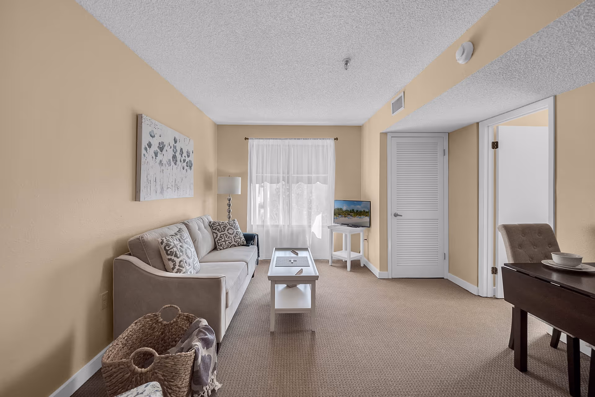 Neutral-toned living room with a sofa, coffee table, TV on a small stand, and a dining area visible through an open doorway.
