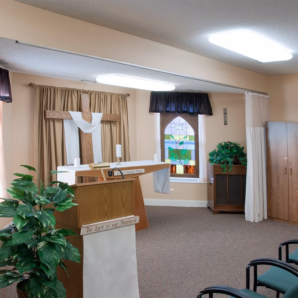 Small chapel with a wooden cross draped in white, an altar and lectern, stained-glass window, and potted plants.