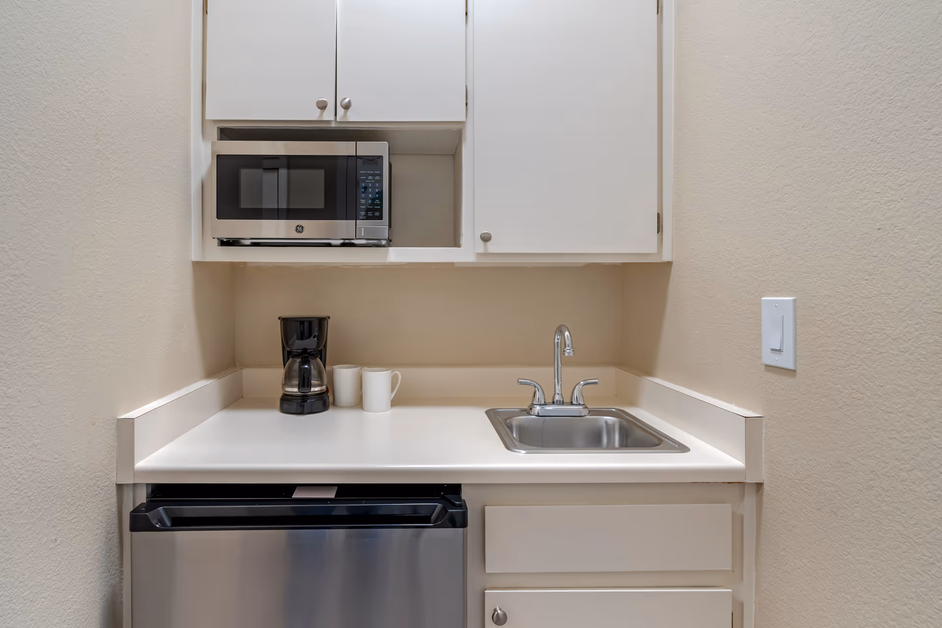 A small kitchenette with white cabinets, a stainless steel microwave, a coffee maker with two white mugs, a stainless steel sink with a faucet, and a mini refrigerator below the counter.