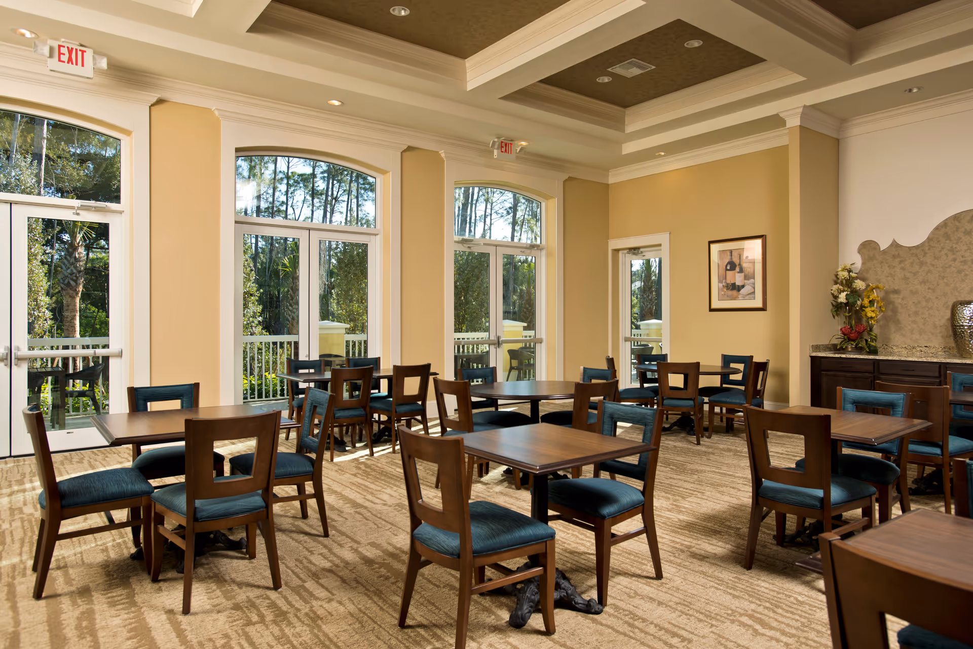 A bright dining room with multiple wooden tables and chairs with blue cushions. Large windows and glass doors let in natural light and offer a view of trees outside. The walls are painted beige with white trim, and there is a framed picture and a floral arrangement on a side counter.