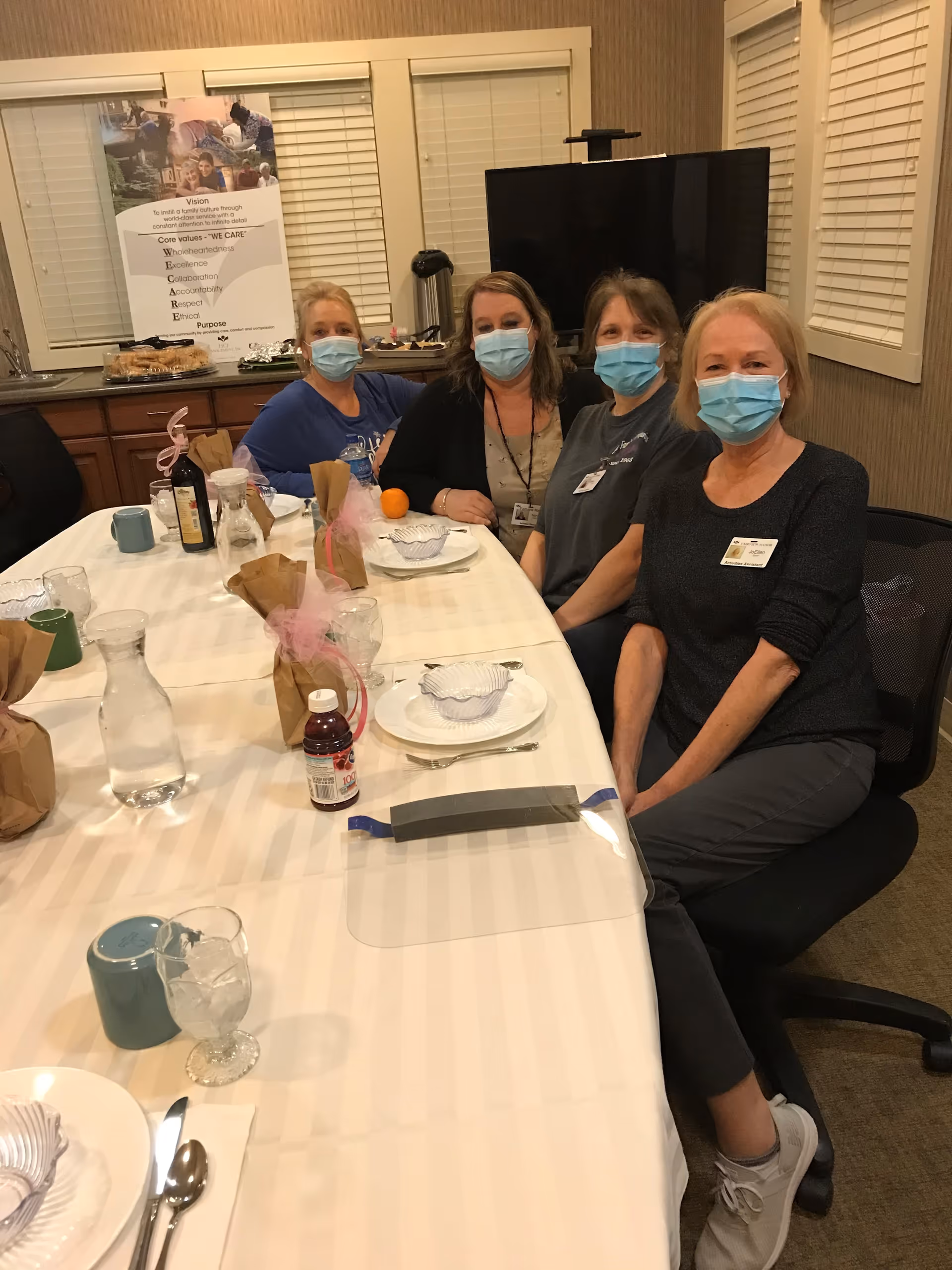 Four masked staff seated at a long table set for a meal in a dining/meeting room of a senior living facility.