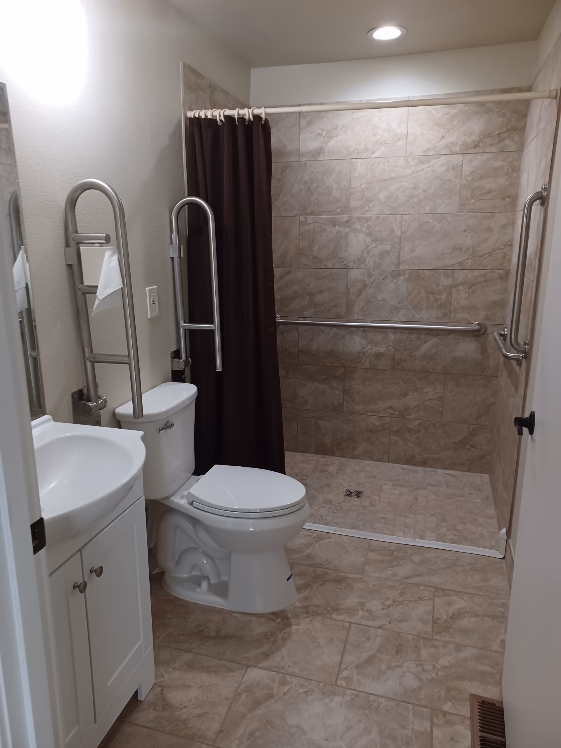 Accessible bathroom with a toilet, sink, grab bars, and a walk-in tiled shower with a brown curtain.