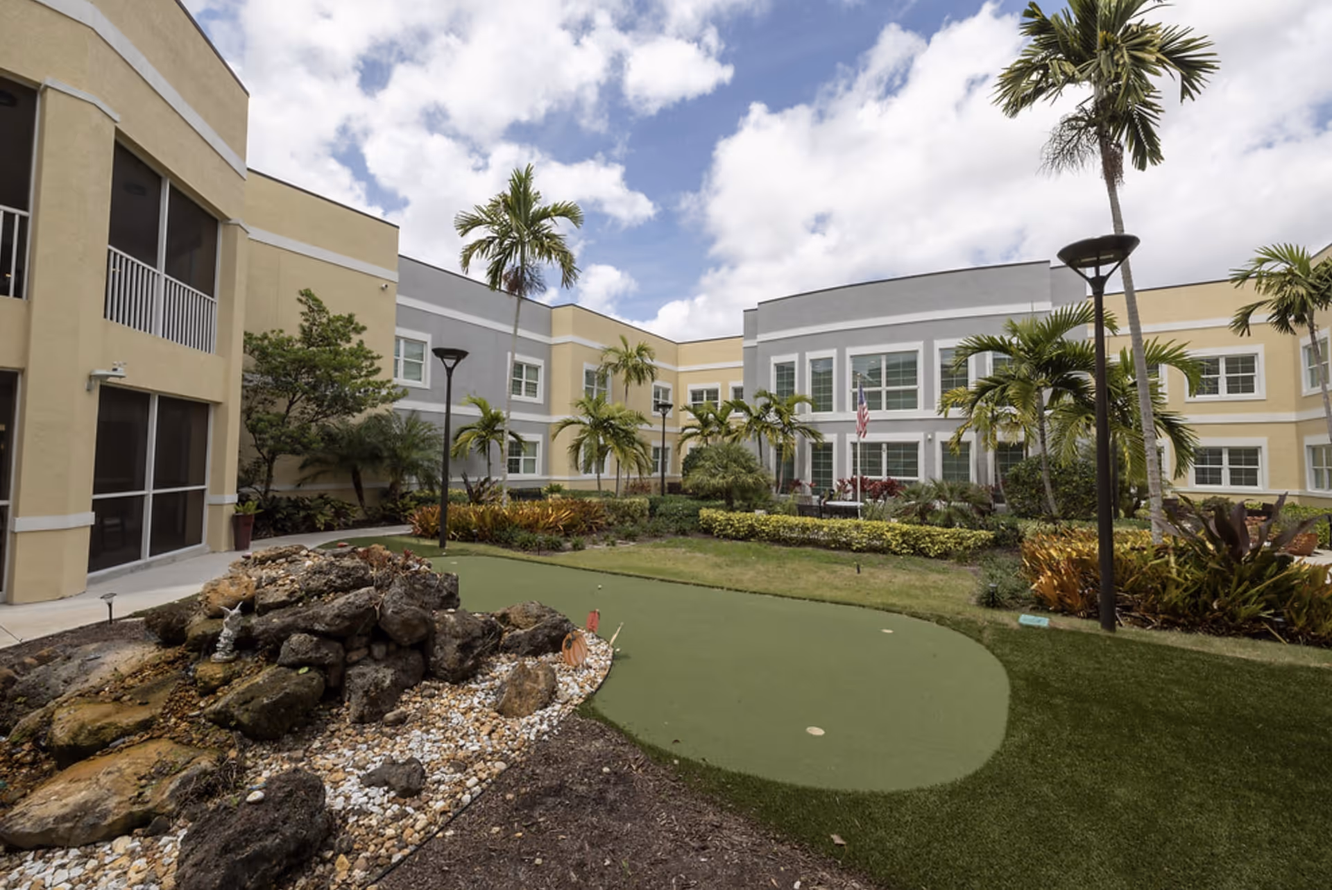 Outdoor courtyard area of a senior living facility with a putting green, landscaped garden with rocks and plants, palm trees, and a two-story building with yellow and gray walls surrounding the courtyard under a partly cloudy sky.